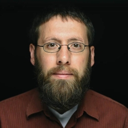 Man with beard and glasses wearing dark brown button up shirt with white t shirt underneath