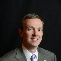 Man with dark grey suit with white dress shirt and purple tie in front of black background