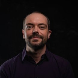 Man with facial hair with dark shirt in front of dark backdrop