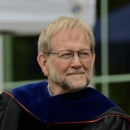 Man with blonde hair and facial hair wearing glasses and PHD graduation gear