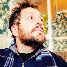 Man with dark hair and beard wearing navy sweater and plaid scarf