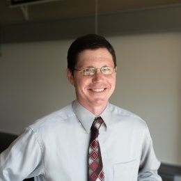 Man with dark hair wearing glasses, grey button up shirt, and red tie