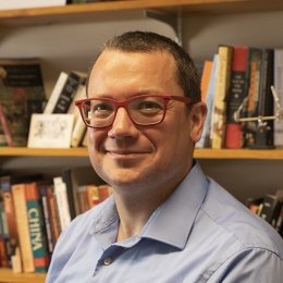 Man with short dark hair wearing red glasses and blue button up dress shirt in front of bookshelf