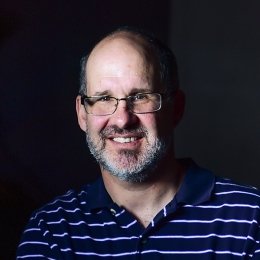 Man with grey beard wearing glasses and navy with white stripes polo in front of black background