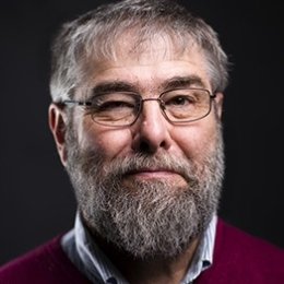 Man with grey hair and beard wearing glasses with red sweater and button up shirt in front of dark background
