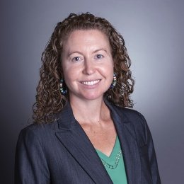 Lady with curly shoulder length brunette hair wearing dark suit jacket with light green sweater in front of grey background
