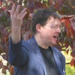 Man gesturing of making a point with hand up in the air wearing sports coat and dark shirt and glasses