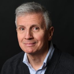 Man with short grey hair wearing black sports coat and blue button up in front of dark studio background