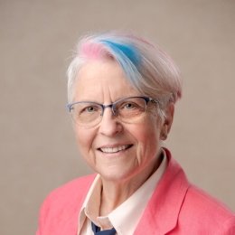 Lady with white hair with painted streaks of blue and pink wearing pink blazer and blue tie with light pink button up dress shirt in front of grey background