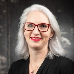 Lady with shoulder length silver hair wearing red glasses, necklace, black top in front of black studio background