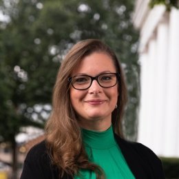 Lady with dark brown hair shoulder length wearing green top and black cardigan with black glasses