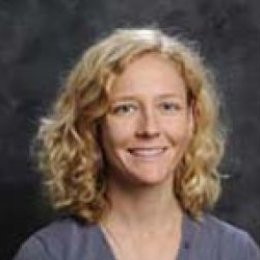 lady with blonde curly hair wearing grey shirt and necklace in front of grey background