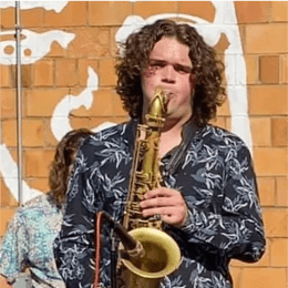 Man with dark curly hair wearing patterned button up shirt playing saxophone