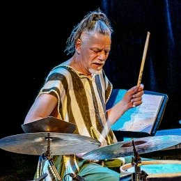 Man with ponytail wearing black and white stripe shirt sitting playing drum set
