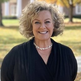 Lady with curly hair with black top and pearls standing on campus in the fall