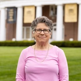 Lady with short curly hair wearing glasses, pink top and necklace in front of old centre