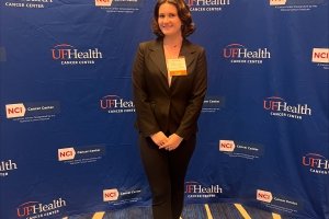 A young woman in a dark business suit poses in front of a conference backdrop with "UF Health Cancer Center" written on it.