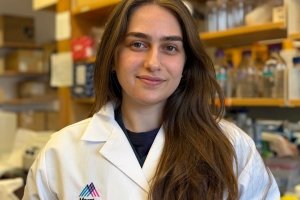 Anna Bostoganashvili is smiling and posing in a lab. Behind her is a shelf of research equipment. She is wearing a lab coat that says Mount Sinai.