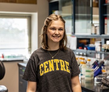 Student standing in lab wearing Centre tshirt and smiling at photographer