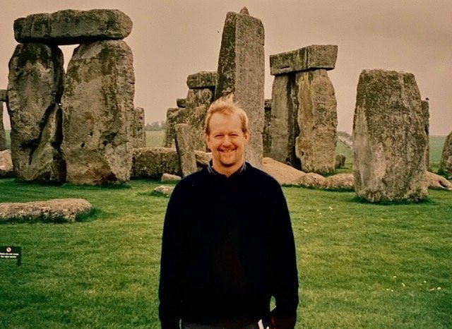 A man in a darm sweater stands in front of Stonehenge, an ancient collection of massive stones arranged in an intricate manor. 