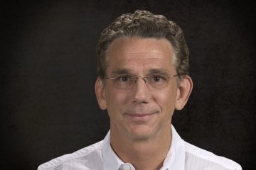 Man with curly hair wearing glasses and white button up shirt in front of dark backdrop