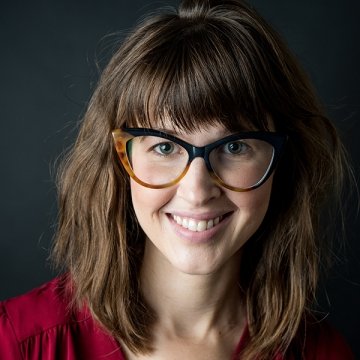 Lady with short brown hair wearing glasses with red top in front of grey background