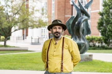 Man with facial hair wearing hat and suspenders and yellow button up shirt standing in front of flame statue