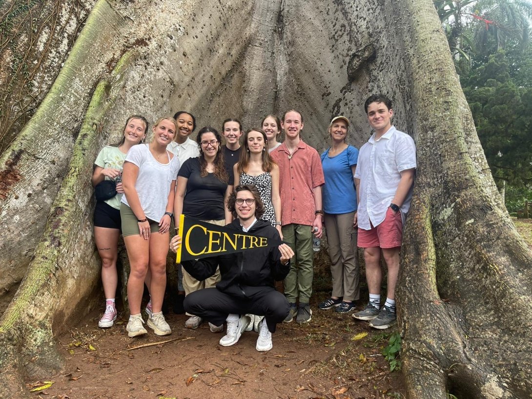 CentreTerm students in Ghana - group of students standing in front of large tree roots. One student holding Centre pennant. 