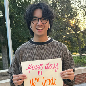 Man with long dark hair wearing glasses, airpods, holding sign first day of 16th grade