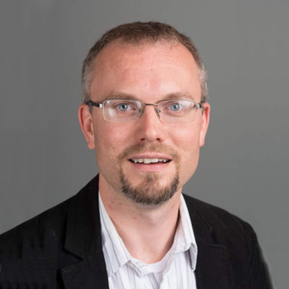 Smiling man with goatee wearing glasses and a blzer against a gray background