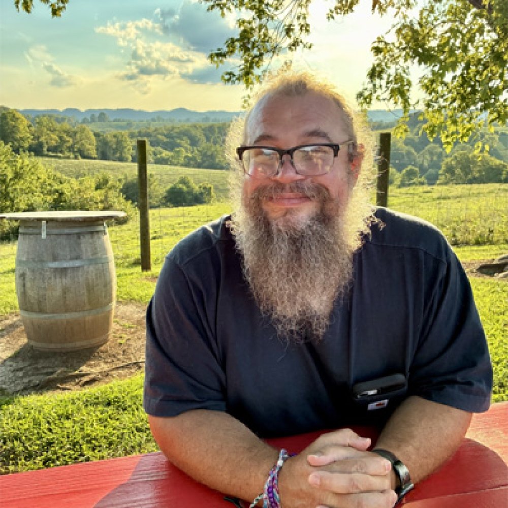 Person wearing black button up shirt while seated at picnic table smiling at photographer