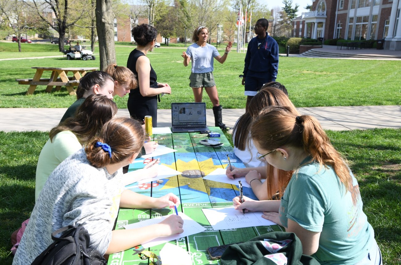 Students participating in discussion at picnic table with laptop and group of students nearby