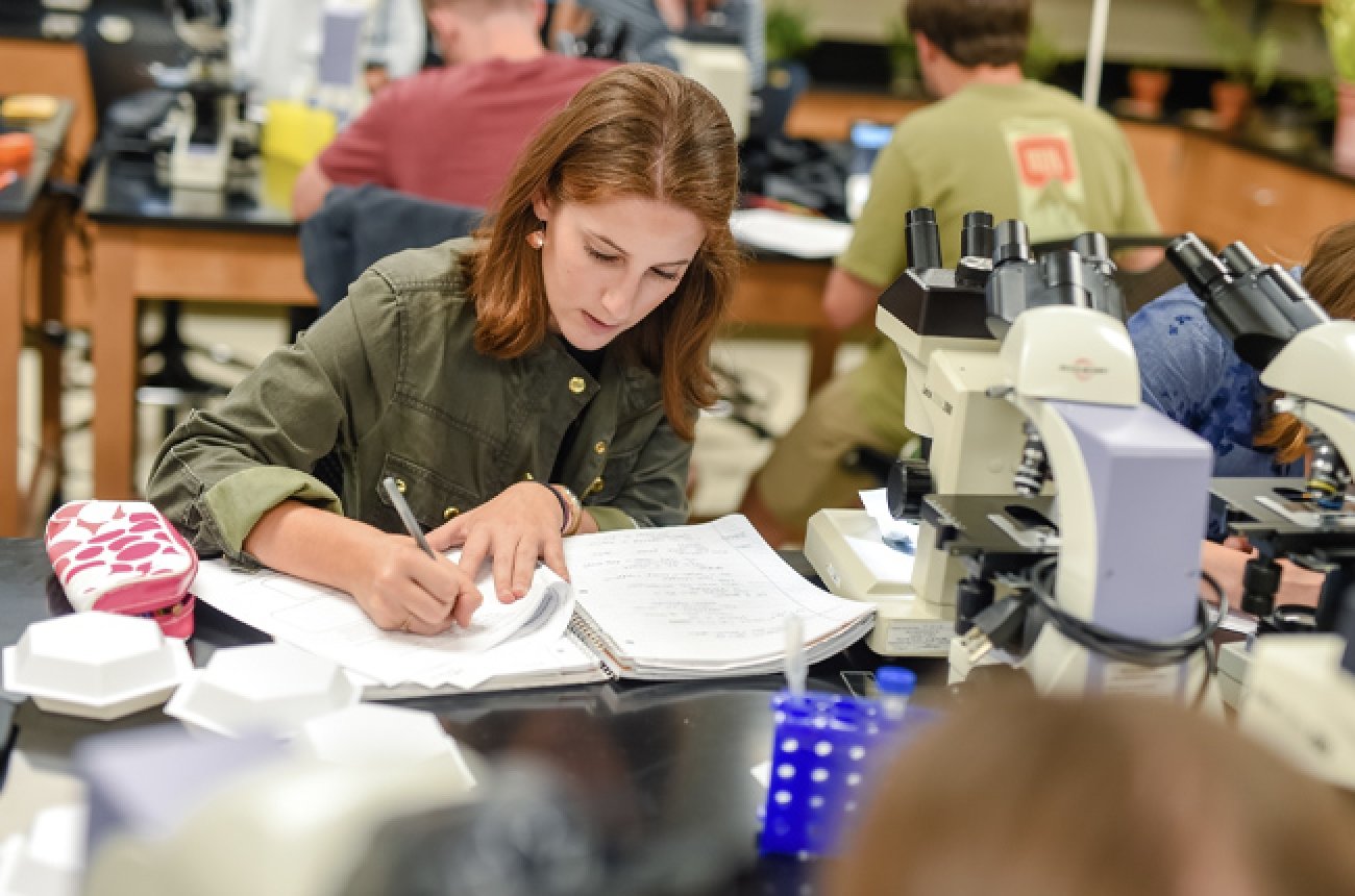 Student in science lab taking notes sitting next to microscope