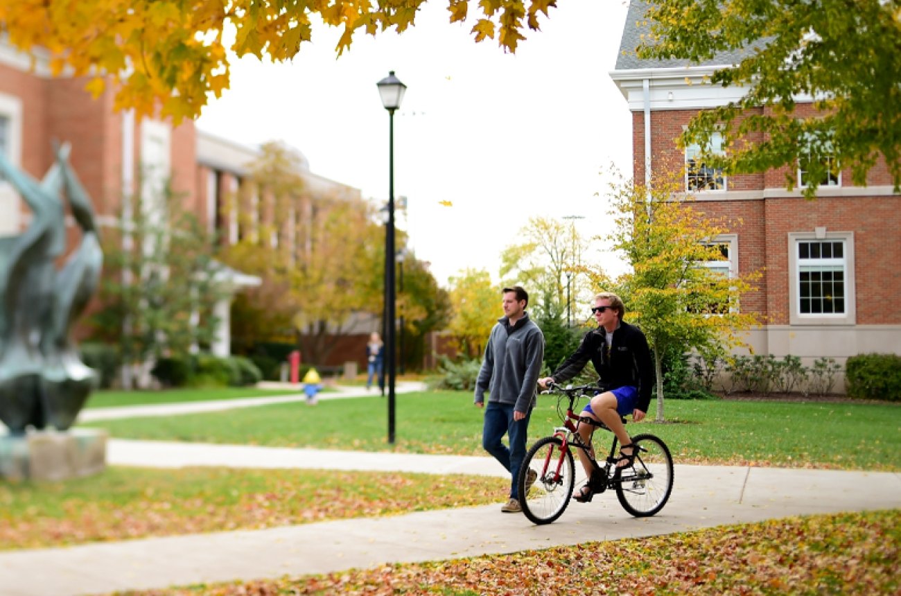 Student riding bike and student walking on campus near the flame statue