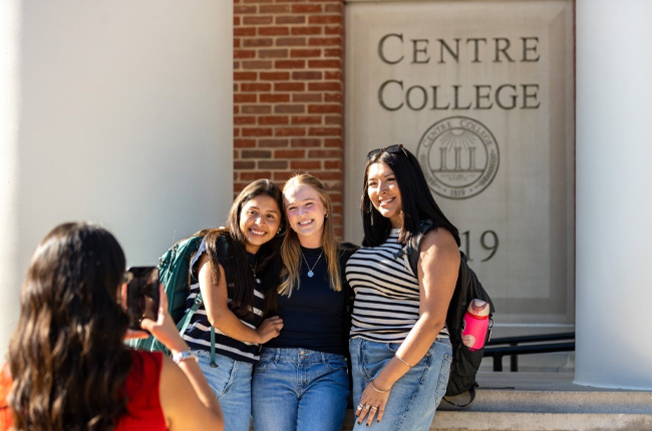 Three students posing for photo in front of library while another student takes photo with phone camera