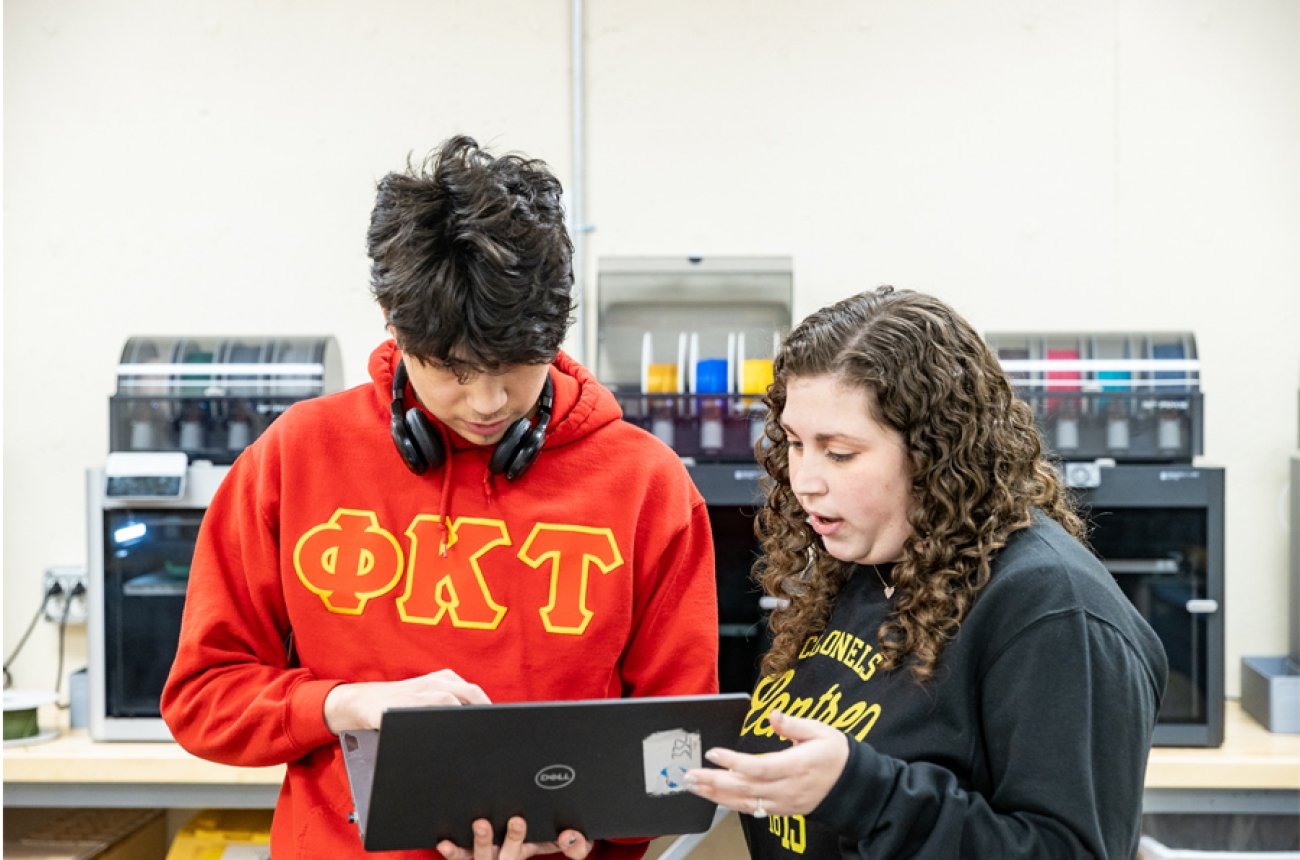 Faculty speaking with student standing in front of 3-d printers