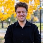Person with brown hair in black sweater standing in front of yellow tree in fall