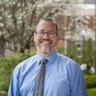 smiling man with graying beard in a blue shirt and tie standing outside