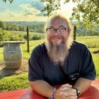 Person wearing black button up shirt while seated at picnic table smiling at photographer