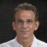 Man with dark curly hair wearing glasses and white button up dress up shirt in front of black background