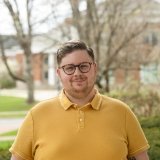 A headshot of Jimmy Schlemmer. He is smiling and wearing a yellow polo shirt, standing in front of campus.