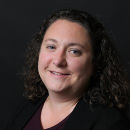 Kerry Paumi headshot, lady with dark curly hair wearing black top with black background