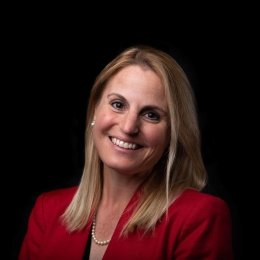 Lady with shoulder length blonde hair with red blazer and pearls in front of black background