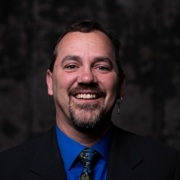 Man with dark hair facial hair with dark suit in front of dark background