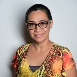 Lady with short dark hair wearing glasses, multi color short sleeve top in front of grey wall