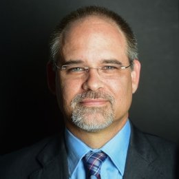 Man with dark hair and gotee wearing glasses, dark suit, light blue dress shirt, and plaid tie in front of grey background