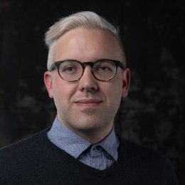 Man with blonde hair wearing glasses with dark sweater and blue button up dress shirt in front of dark background