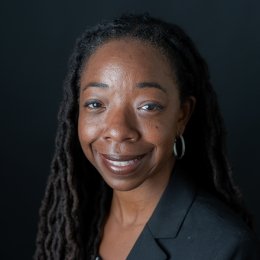 Lady with dark hair wearing black blazer and hoop earrings in front of black background