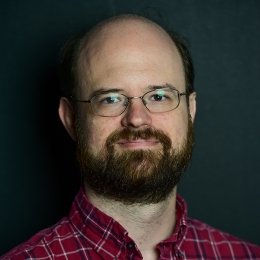 Man with beard wearing glasses and red plaid button up shirt in front of dark background