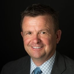 Man with dark hair wearing dark suit with plaid dress shirt and blue tie